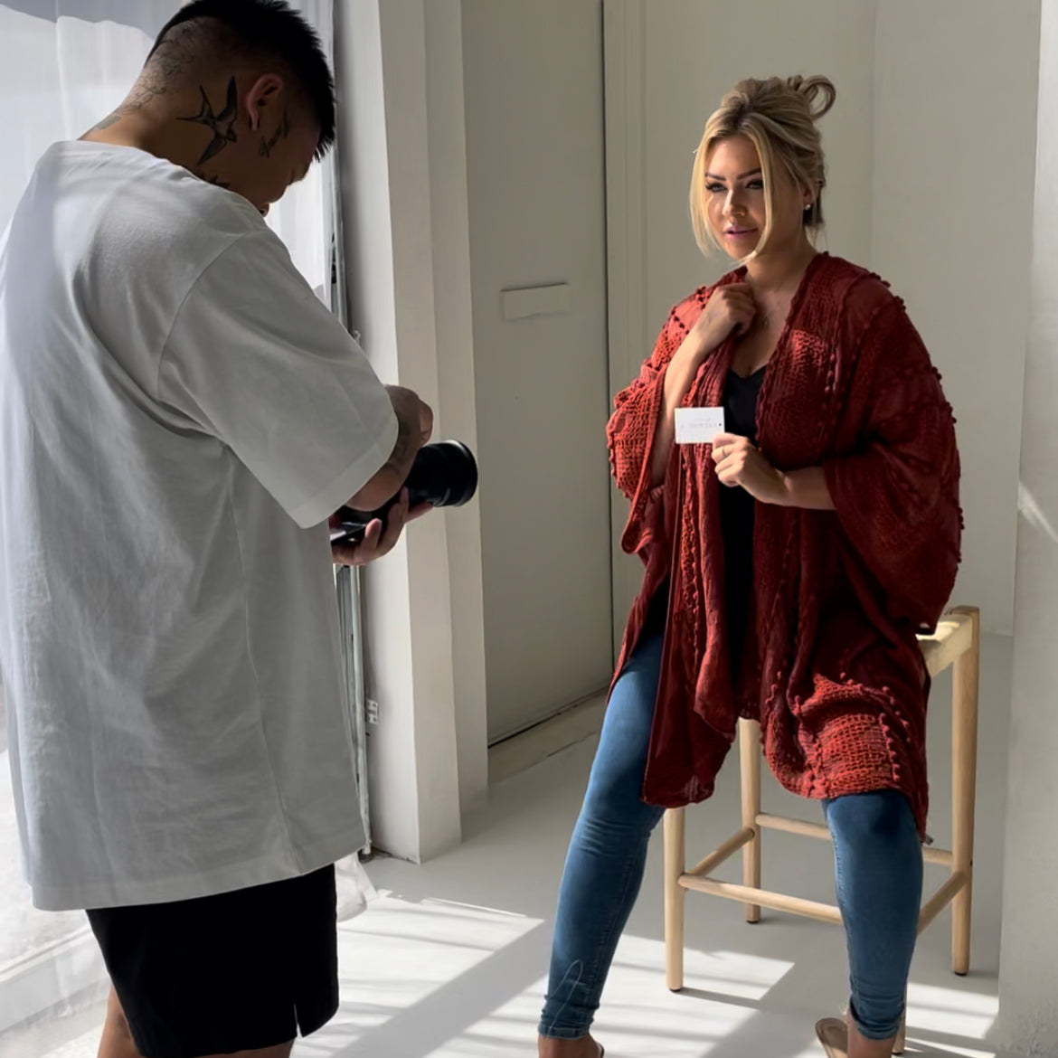 Woman sitting on stool wearing orange sweater and jeans with sandal heels staring at photographer while photographer is taking photo and sun is shining in on them,T.K.S