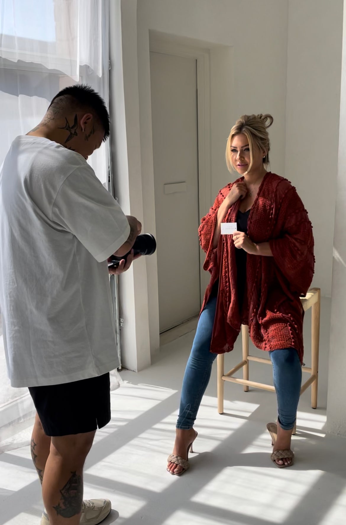 Woman sitting on stool wearing orange sweater and jeans with sandal heels staring at photographer while photographer is taking photo and sun is shining in on them,T.K.S