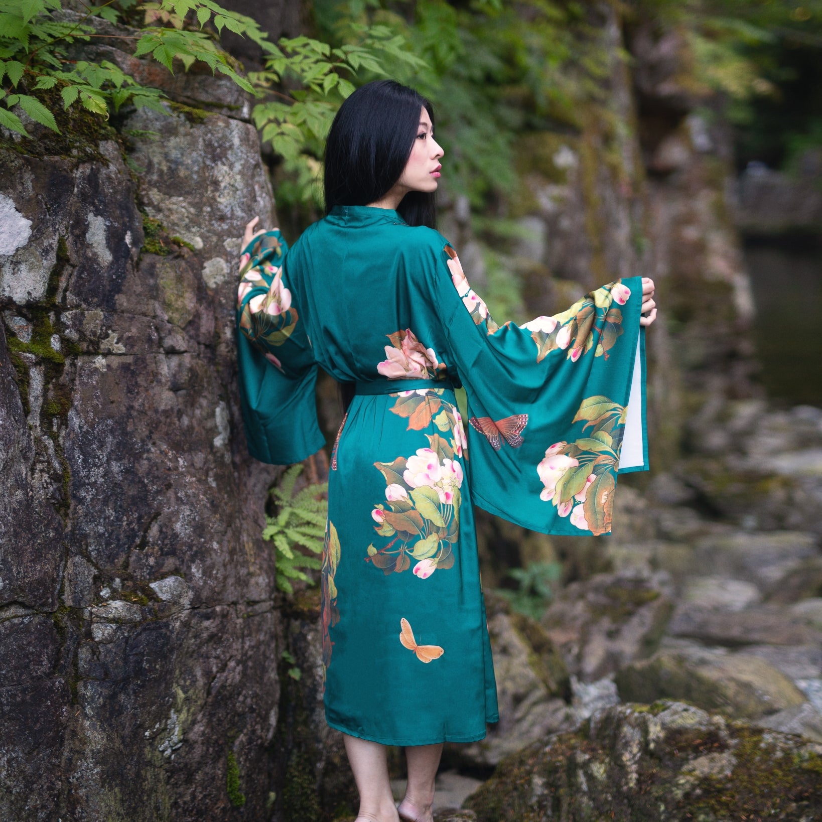 Woman standing beside mossy rock wearing long green floral Jasmine Robe looking away from the camera and into the distance, Shop T.K.S