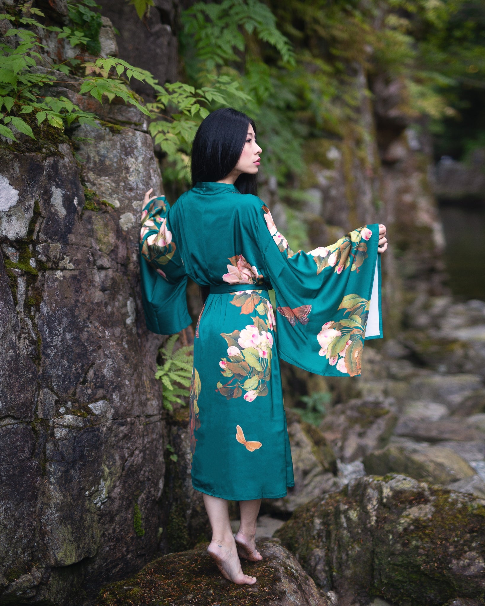 Woman standing beside mossy rock wearing long green floral Jasmine Robe looking away from the camera and into the distance, Shop T.K.S