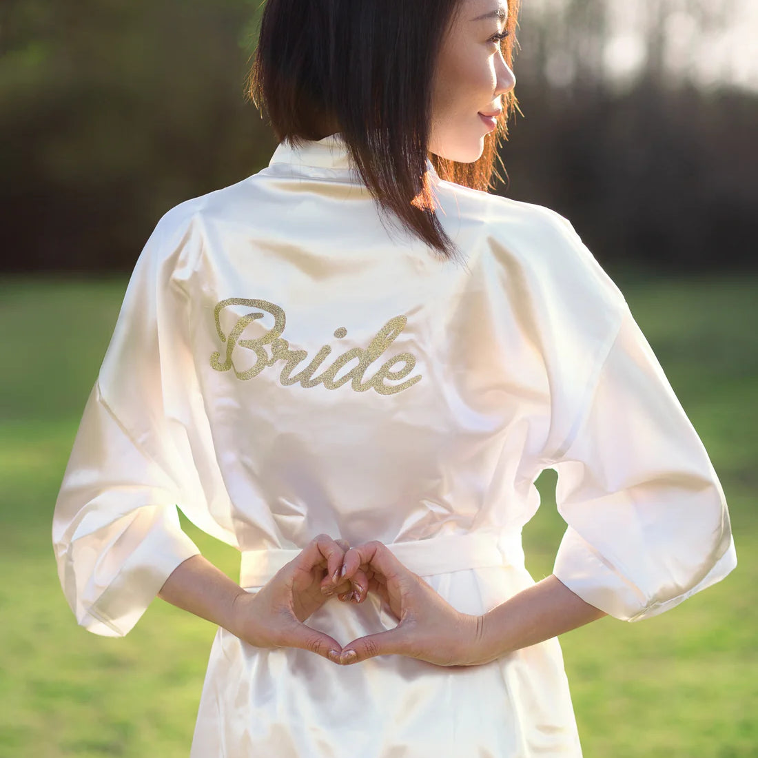 Woman wearing white robe with gold bride lettering on it looking to the side making a heart with her hands on her back, Shop T.K.S