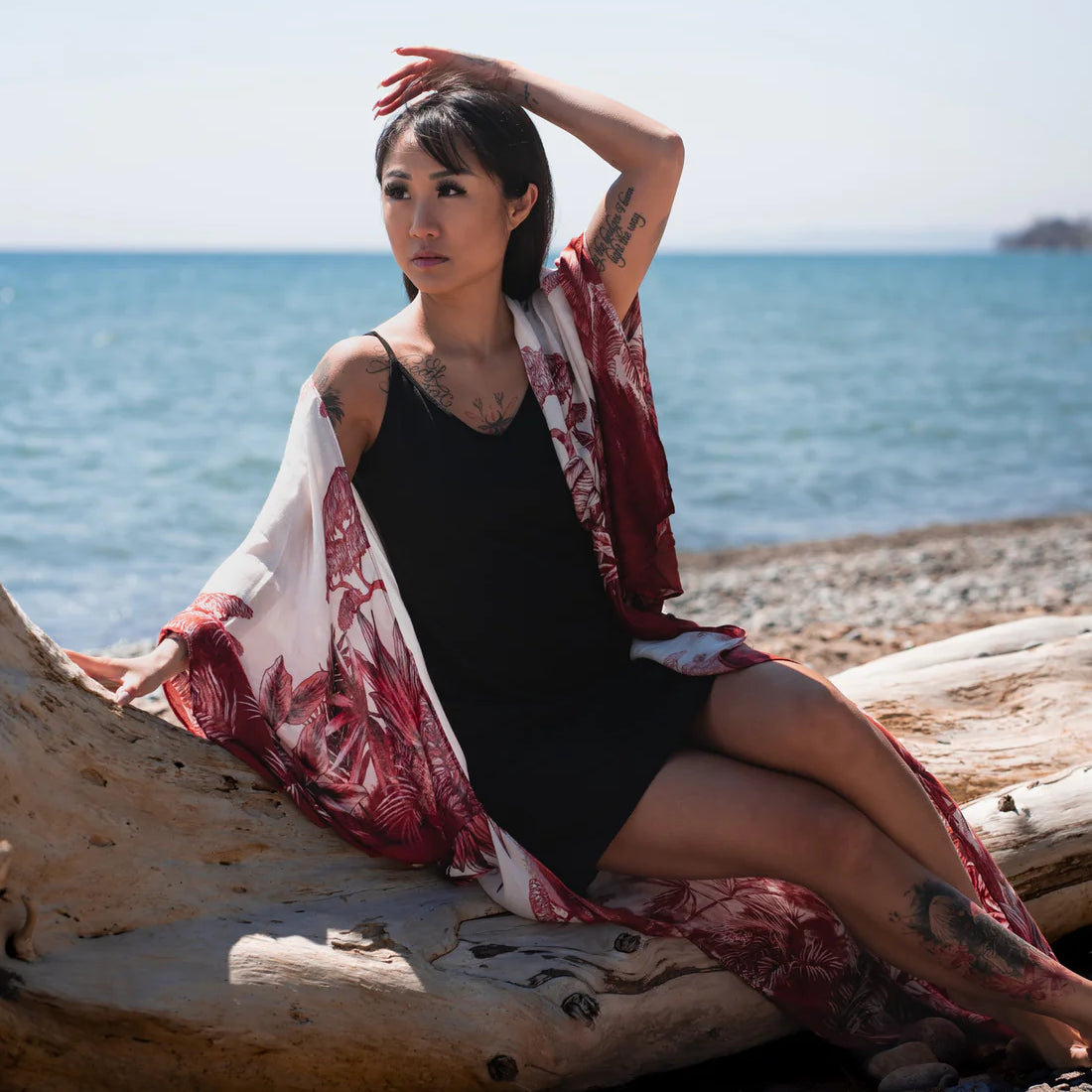 Woman sitting on thick tree branch with one hand in her hair staring away from camera wearing black spaghetti strap bamboo dress and long white and red floral beach boho kimono cover up, Shop T.K.S