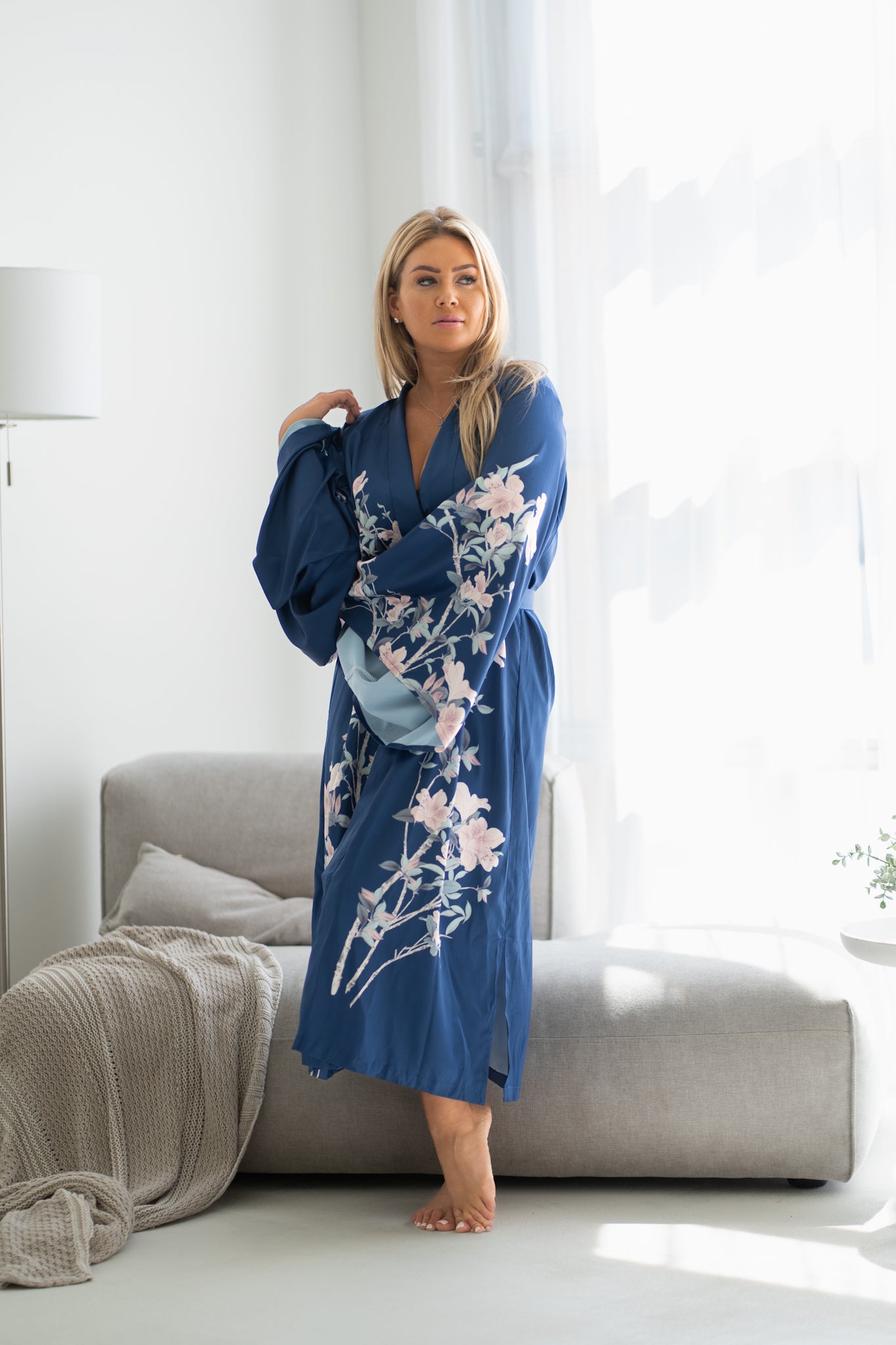 Woman wearing long navy blue floral robe with belt standing near couch, Shop TKS
