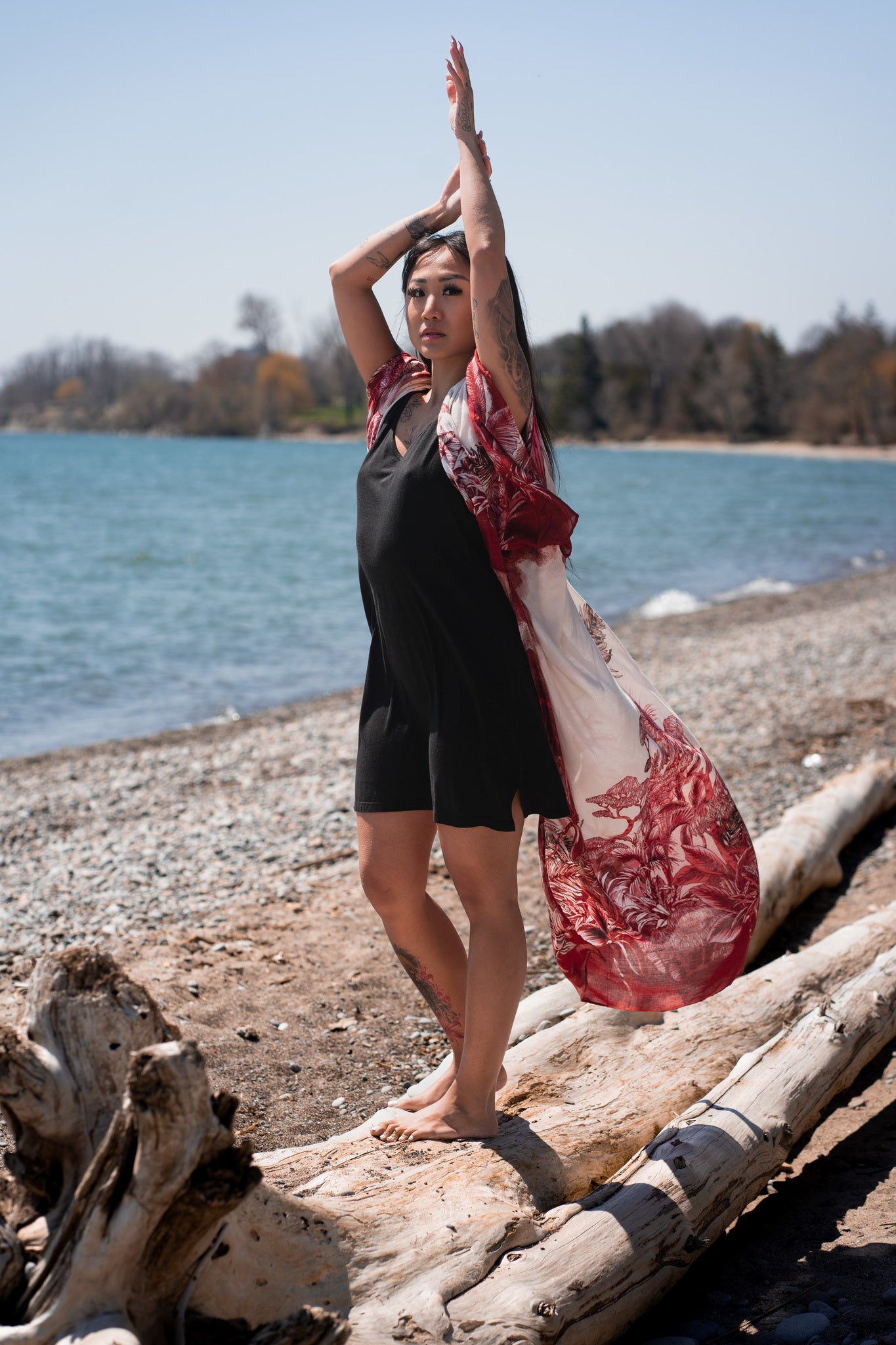 woman standing on log with hands up wearing long flowy white beach over up and black dress