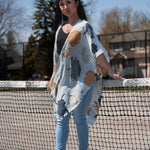 Woman wearing white floral beach cover up standing in front of tennis court wearing black bamboo tank and jeans and sandals