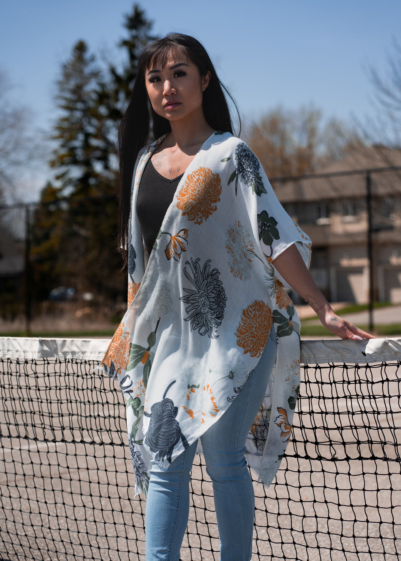 Woman wearing white floral beach cover up standing in front of tennis court net, with jeans and bamboo tank top, Shop T.K.S