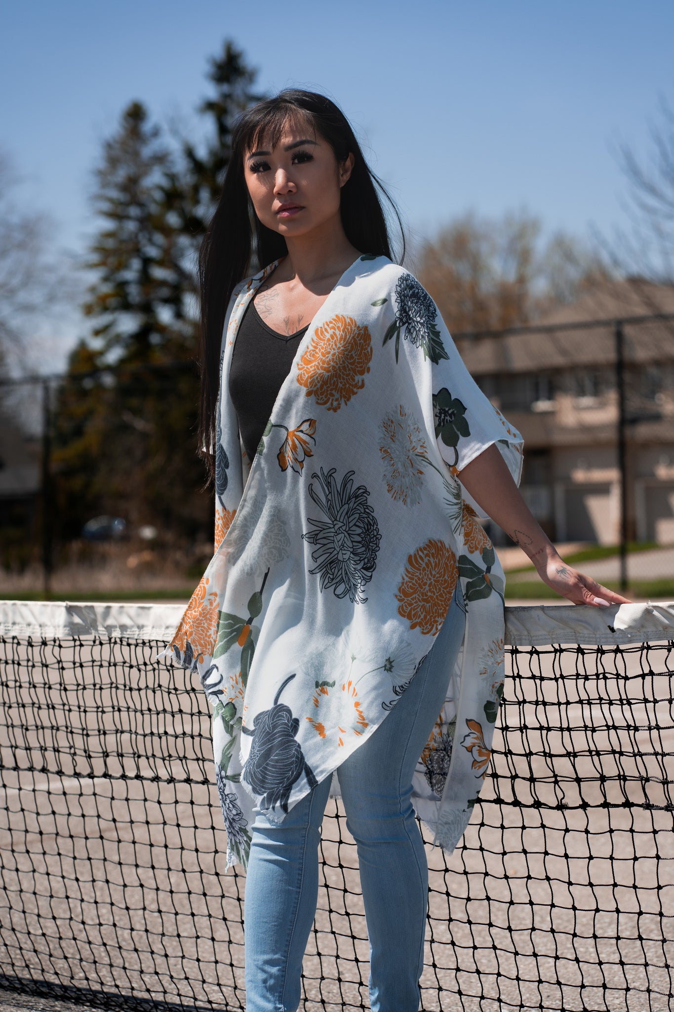 Woman wearing white floral beach cover up standing in front of tennis court net, with jeans and bamboo tank top, Shop T.K.S
