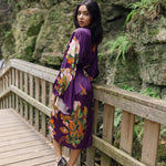 Women's long robe with purple floral and butterfly design, women looking over shoulder wearing ballet flats, with cliff rock hiking and mountains in background