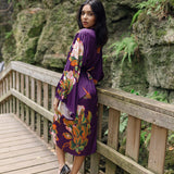 Women's long robe with purple floral and butterfly design, women looking over shoulder wearing ballet flats, with cliff rock hiking and mountains in background