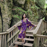 Woman looking over shoulder standing on bridge wearing long purple robe with kimono sleeves and butterfly and floral print design