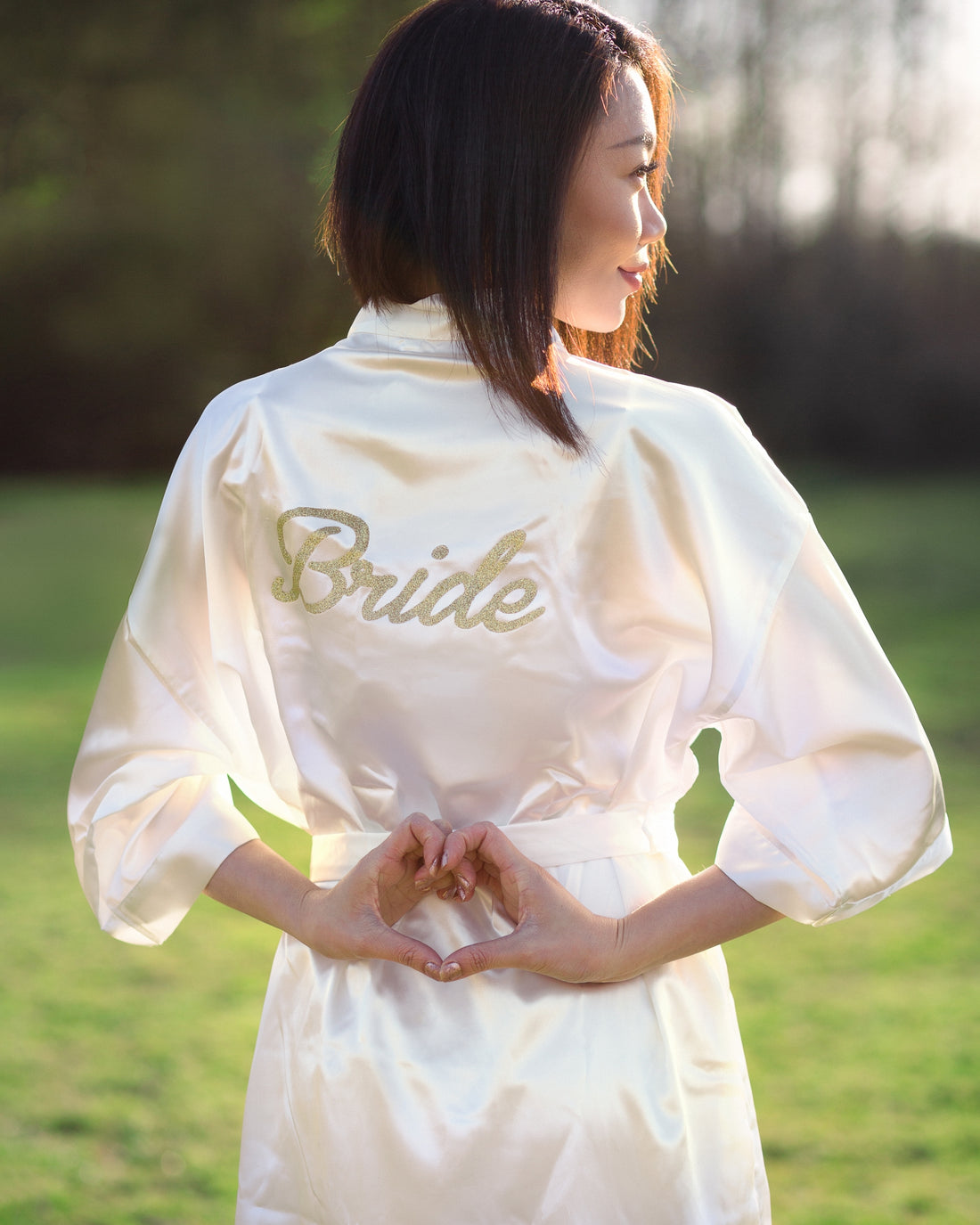 woman standing staring to the side wearing white robe that says bride in gold letters making a heart with her hands, Shop T.K.S