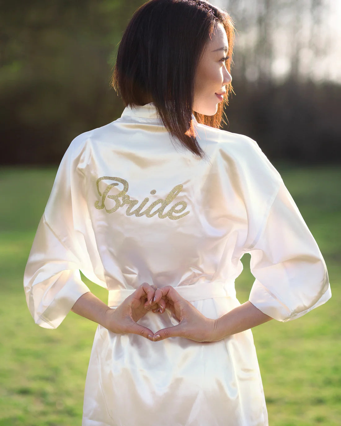 Woman wearing white robe with gold bride lettering on it looking to the side making a heart with her hands on her back, Shop T.K.S