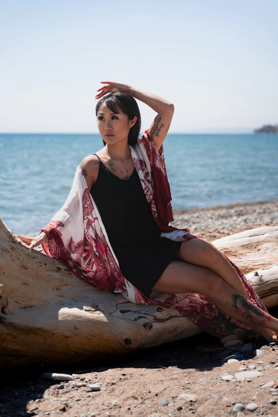 Woman sitting on thick tree branch with one hand in her hair staring away from camera wearing black spaghetti strap bamboo dress and long white and red floral beach boho kimono cover up, Shop T.K.S