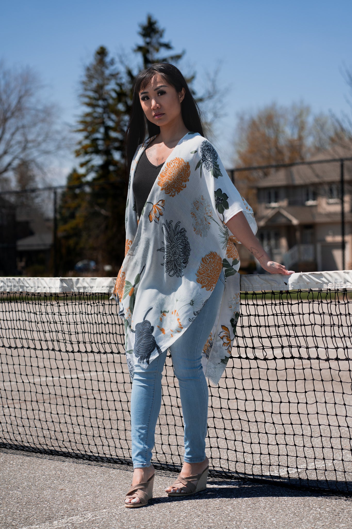Woman wearing white floral beach cover up standing in front of tennis court wearing black bamboo tank and jeans and sandals