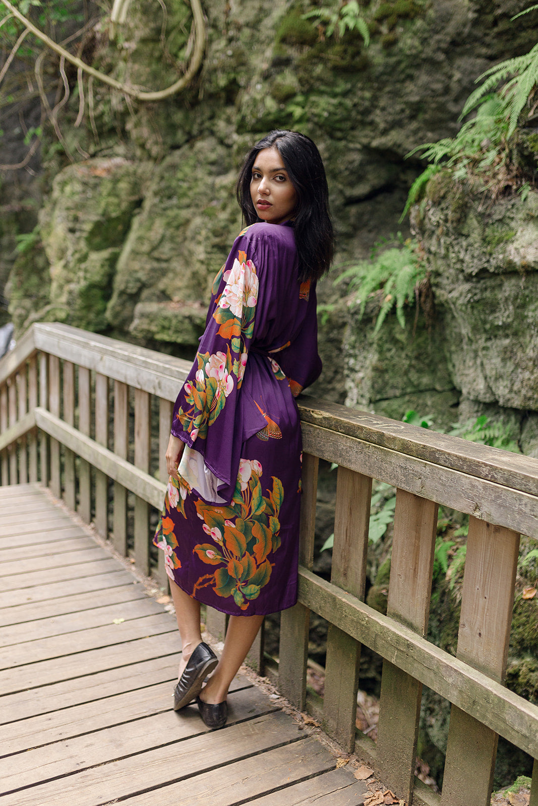 Women's long robe with purple floral and butterfly design, women looking over shoulder wearing ballet flats, with cliff rock hiking and mountains in background