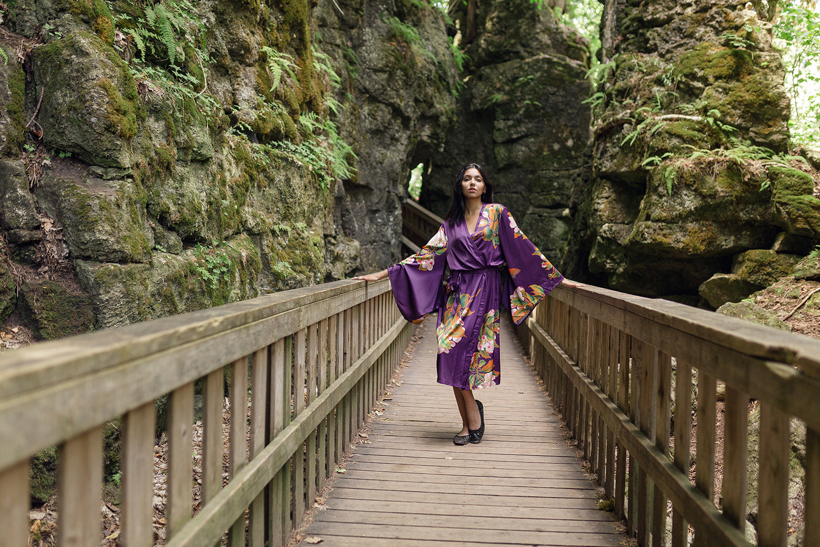 Women standing in between two rocks wearing long floral robe iwth kimono robes and floral and butterfly design, standing on bridge wearing black ballet flats