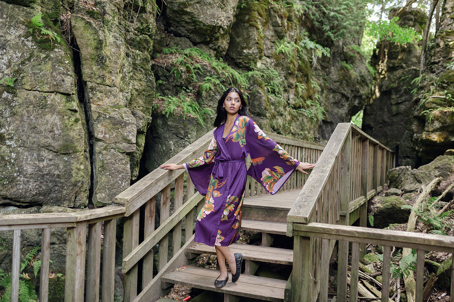Woman looking over shoulder standing on bridge wearing long purple robe with kimono sleeves and butterfly and floral print design