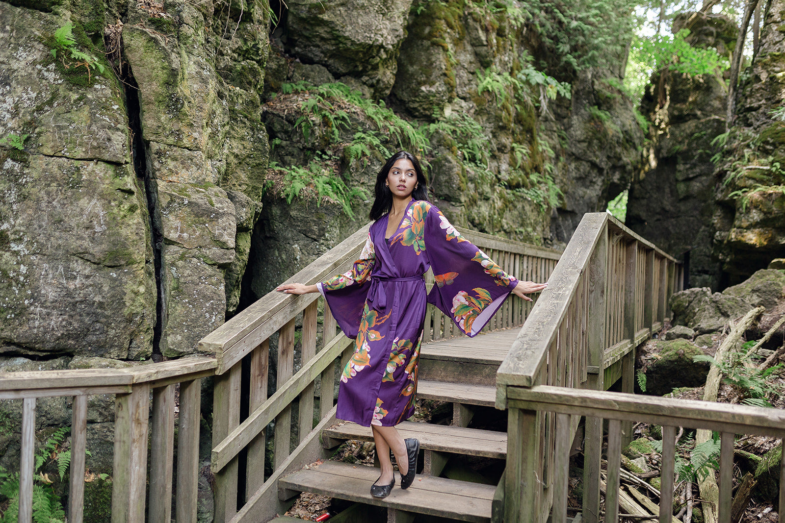 Woman looking over shoulder standing on bridge wearing long purple robe with kimono sleeves and butterfly and floral print design